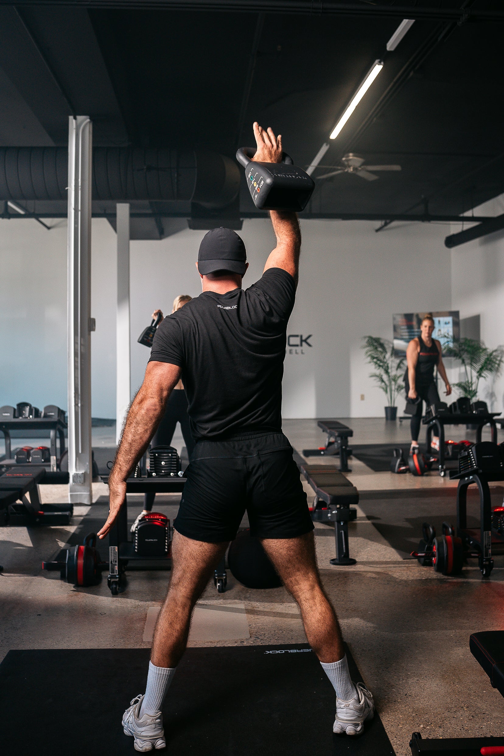 An action shot of a powerblock fitness studio coach teaching from the front of their hiit based class, demonstrating kettlebell shoulder presses.