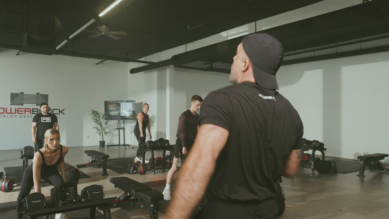 Load video: PowerBlock Fitness Studio video shoot, a coach leading a class through different movements such as kettlebell shoulder presses, jumping jacks, and med ball slams.